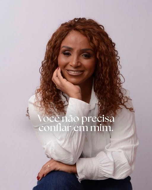 Andréa Maia, de cabelo cacheado ruivo e camisa branca, com a mão no queixo, sorrindo.